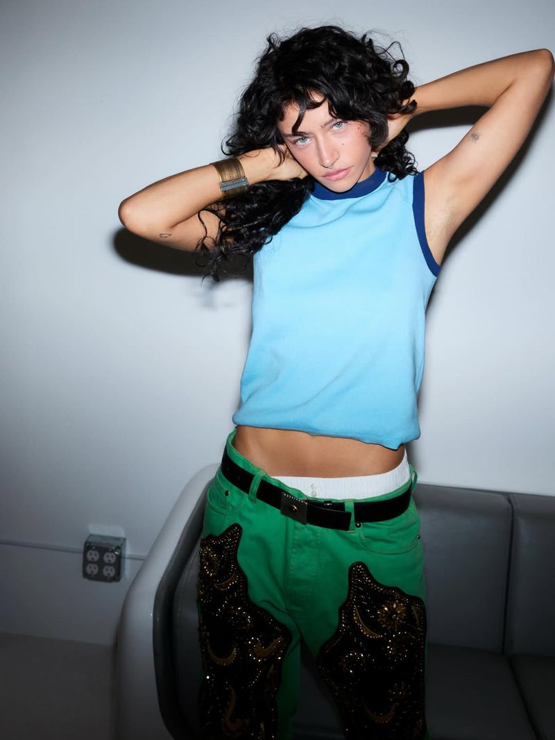 Young adult woman in a blue top and green pants poses indoors with hands behind her head, showcasing a casual and stylish fashion look.