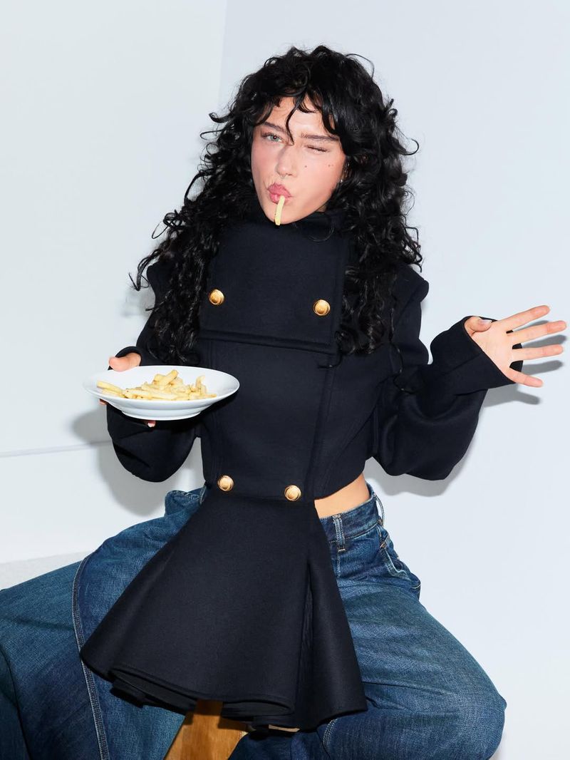Actor Odessa A'zion holding a plate of fries and winking, wearing a black jacket and blue jeans against a white background.
