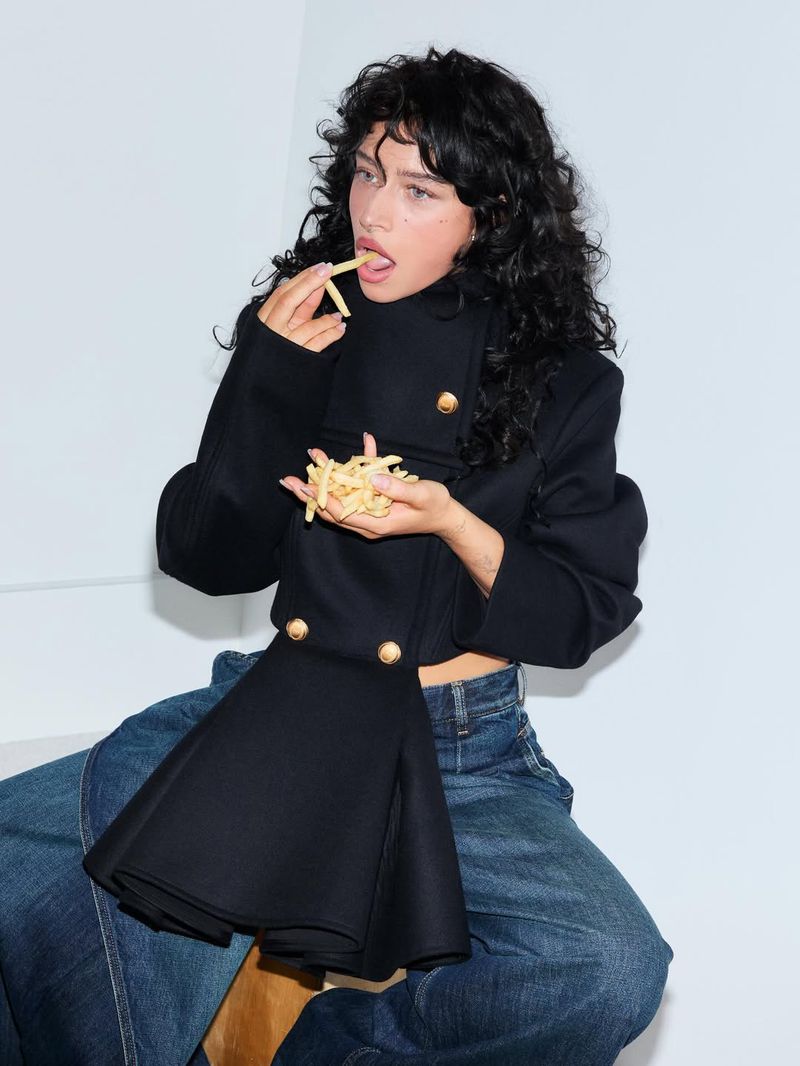 Young woman with curly hair eating French fries while sitting, wearing a navy jacket and wide-leg denim jeans against a white backdrop.