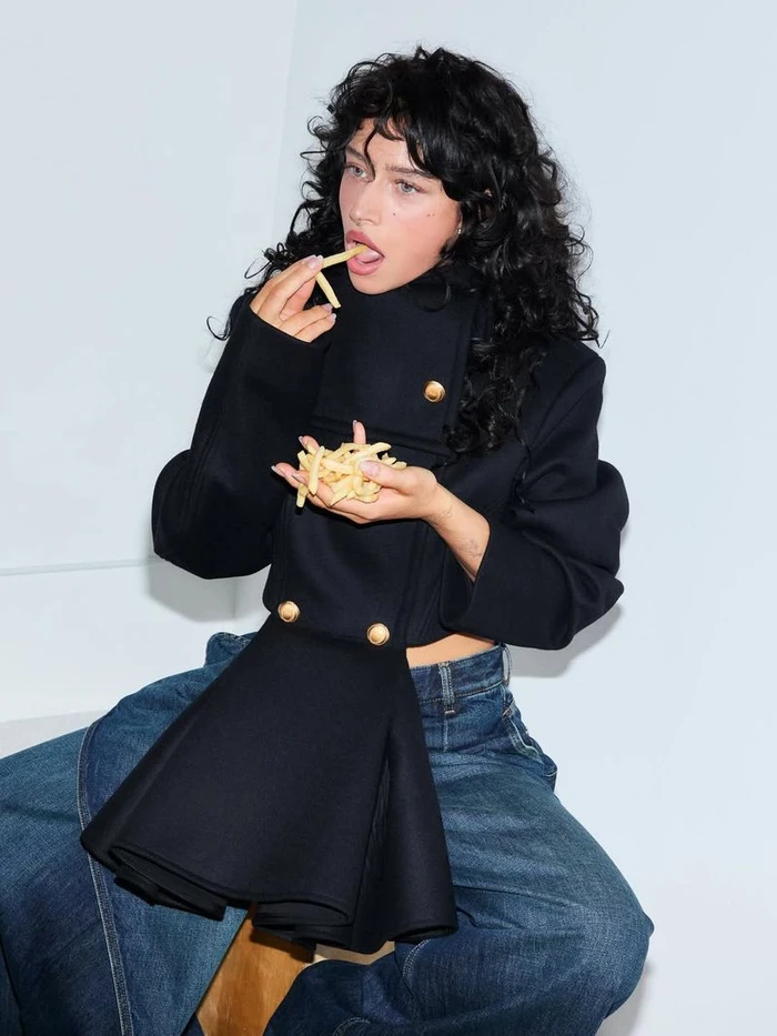 Odessa A'zion wearing a black structured coat and blue jeans eating french fries against a plain white studio background.