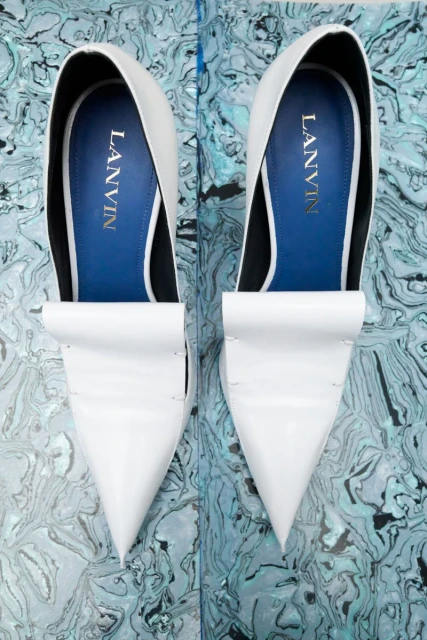 Elevated view of two white Lanvin pointed-toe pumps with blue lining, showcased against a blue and white marbled backdrop.