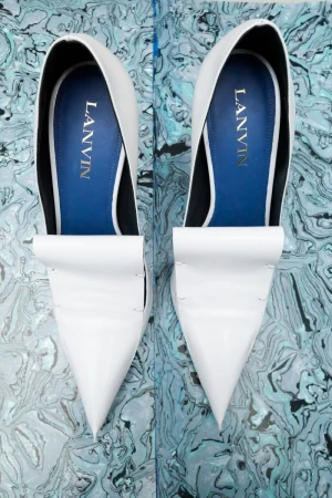 Elevated view of two white Lanvin pointed-toe pumps with blue lining, showcased against a blue and white marbled backdrop.