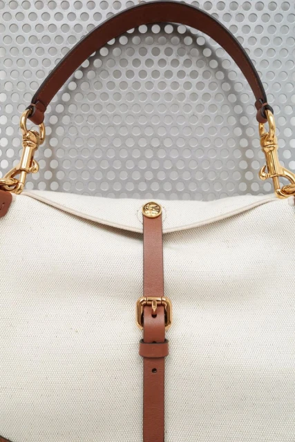 Close-up of a cream-colored canvas handbag with brown leather straps and gold hardware against a textured metal background.