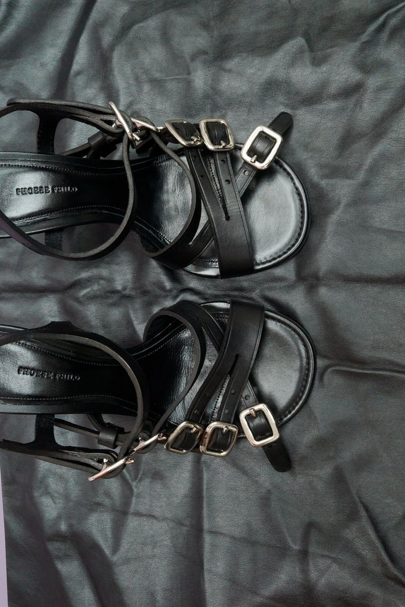 Close-up of a pair of black leather strappy sandals with silver buckles on a black draped fabric background, product photography.