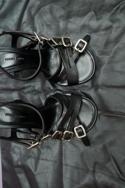 Close-up of a pair of black leather strappy sandals with silver buckles on a black draped fabric background, product photography.