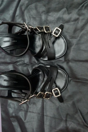 Close-up of a pair of black leather strappy sandals with silver buckles on a black draped fabric background, product photography.