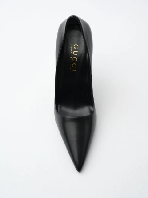 Black leather Gucci stiletto heel on white. The designer shoe is displayed from above showing pointed toe, heel, and gold logo.