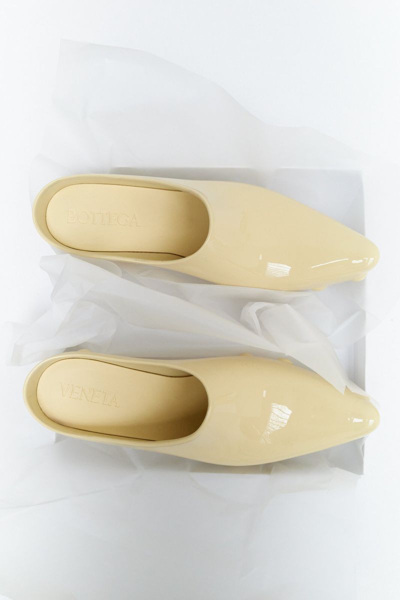 Elevated studio shot of a pair of beige patent-leather heeled slip-on shoes resting on tissue paper, casting soft shadows on a white backdrop.