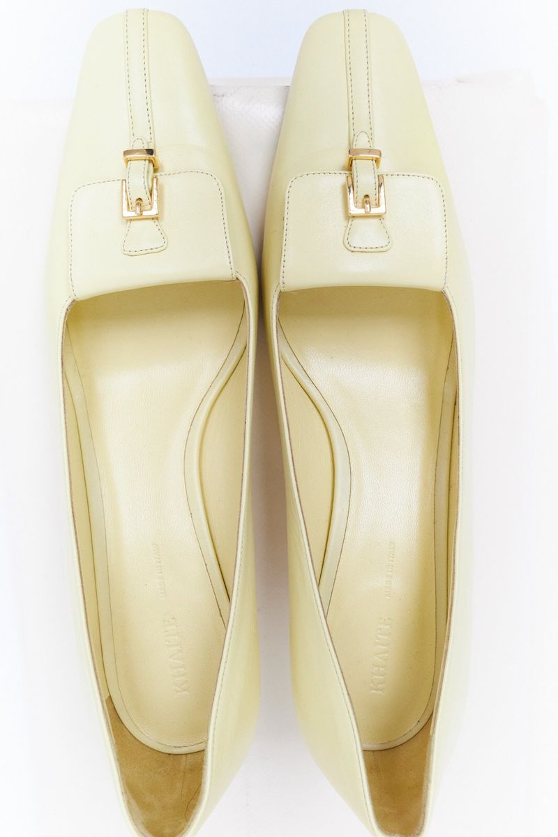 Overhead shot of pair of cream-colored leather heeled shoes with gold buckle detail against a white background. Stylish women's footwear.