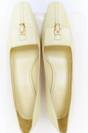 Overhead shot of pair of cream-colored leather heeled shoes with gold buckle detail against a white background. Stylish women's footwear.