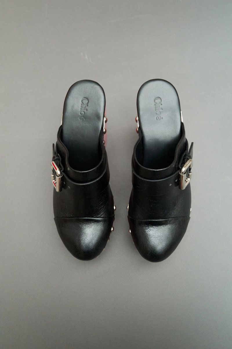 Overhead shot of a pair of black leather clog shoes with decorative silver buckles and embellishments on a gray background.