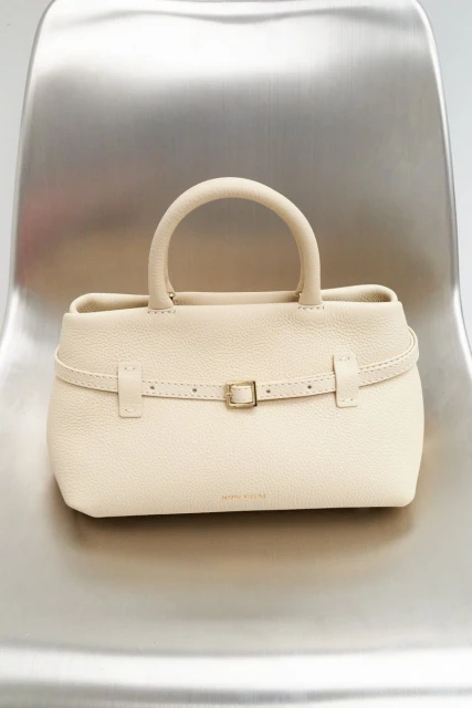 Close-up studio shot of a cream-colored leather handbag with handle and belt detail, placed on a modern silver metal chair.