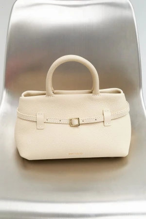 Close-up studio shot of a cream-colored leather handbag with handle and belt detail, placed on a modern silver metal chair.