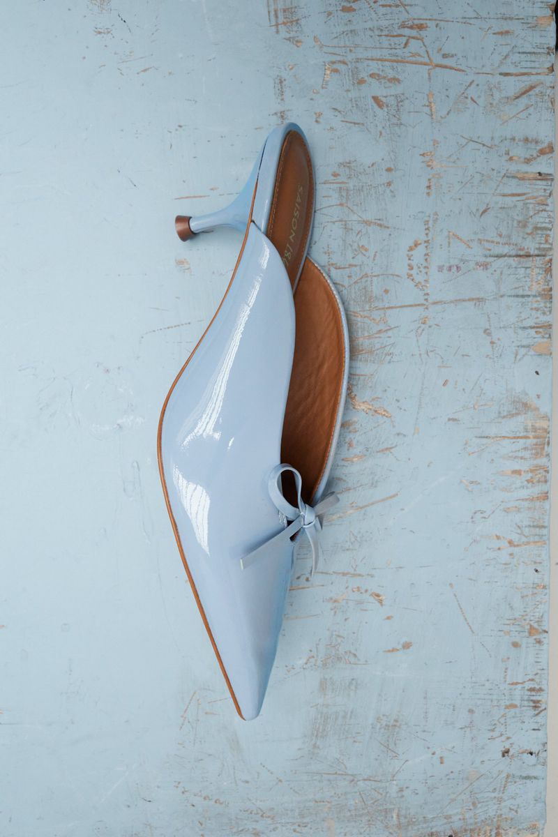 Light blue heeled mules with a pointed toe and small bow detail, set against a light blue surface with rustic texture.