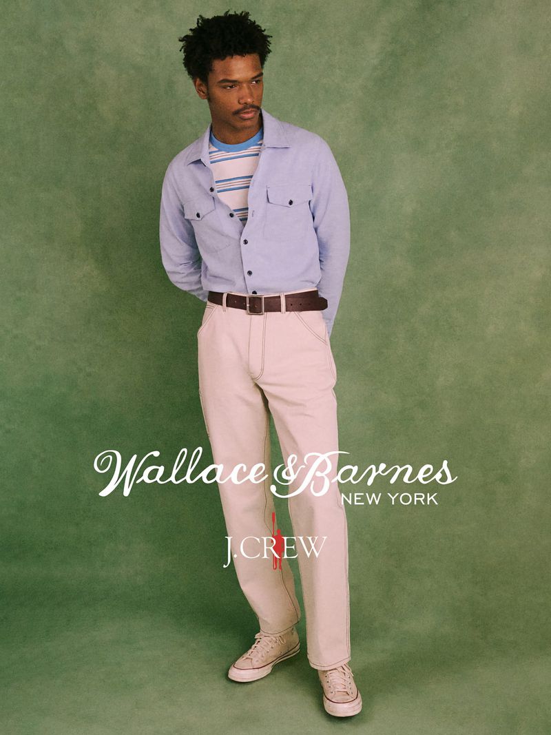 A young adult male poses in a light blue button-down shirt, beige pants, and beige sneakers against a textured green backdrop indoors.