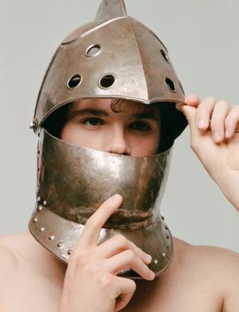 Man wears ornate, aged metal helmet and touches the visor with his hand in a close-up studio shot on a neutral background.