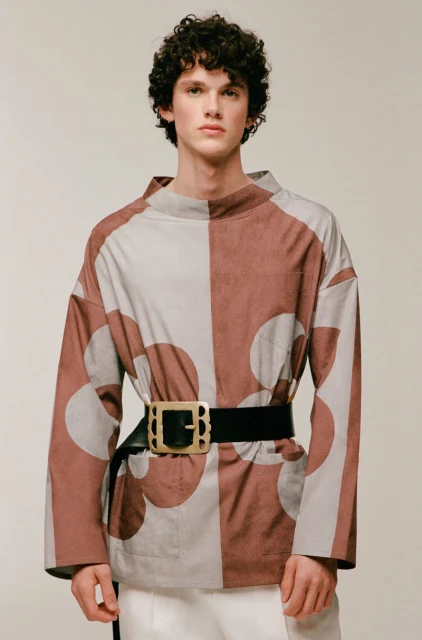 A young adult male poses in a studio wearing a brown and beige geometric print shirt, white pants, and a black belt with a gold buckle.