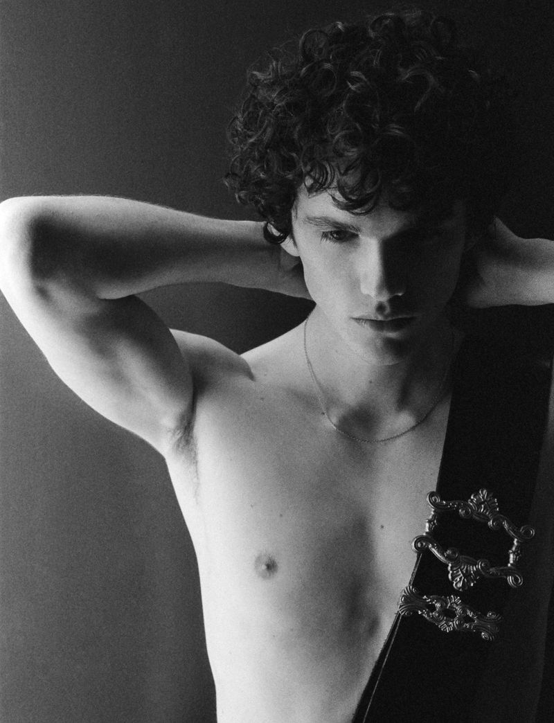 Black and white portrait of a curly-haired young man with bare torso wearing a decorative sash, posing against a dark backdrop.