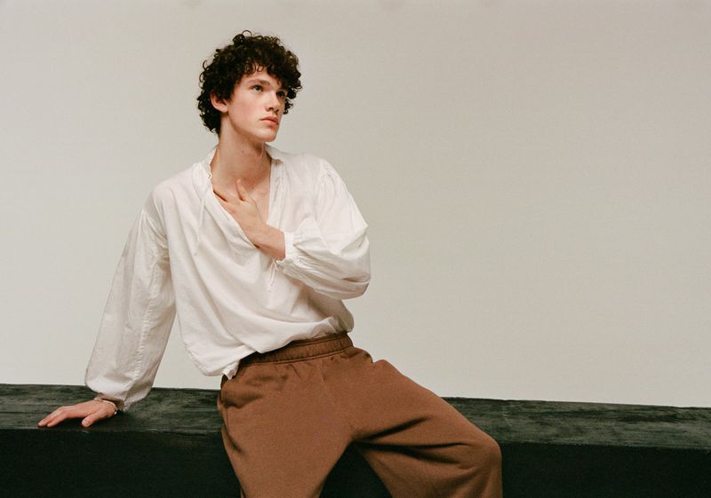 Young adult man with curly hair poses in a white, long-sleeved shirt and brown pants against a neutral backdrop, his hand gently touching his chest.