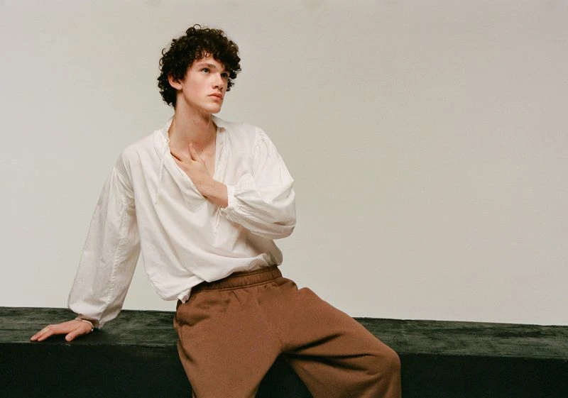Young adult man with curly hair poses in a white, long-sleeved shirt and brown pants against a neutral backdrop, his hand gently touching his chest.