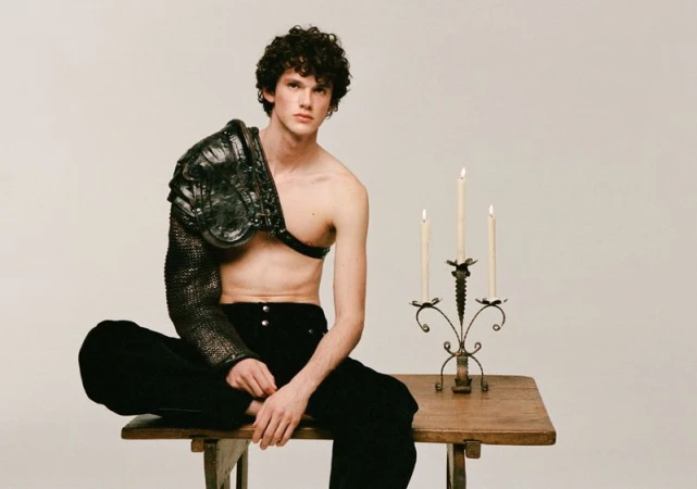 Young adult male poses on a wooden table indoors, wearing modern black pants and ornate metal shoulder armor with lit candles nearby.