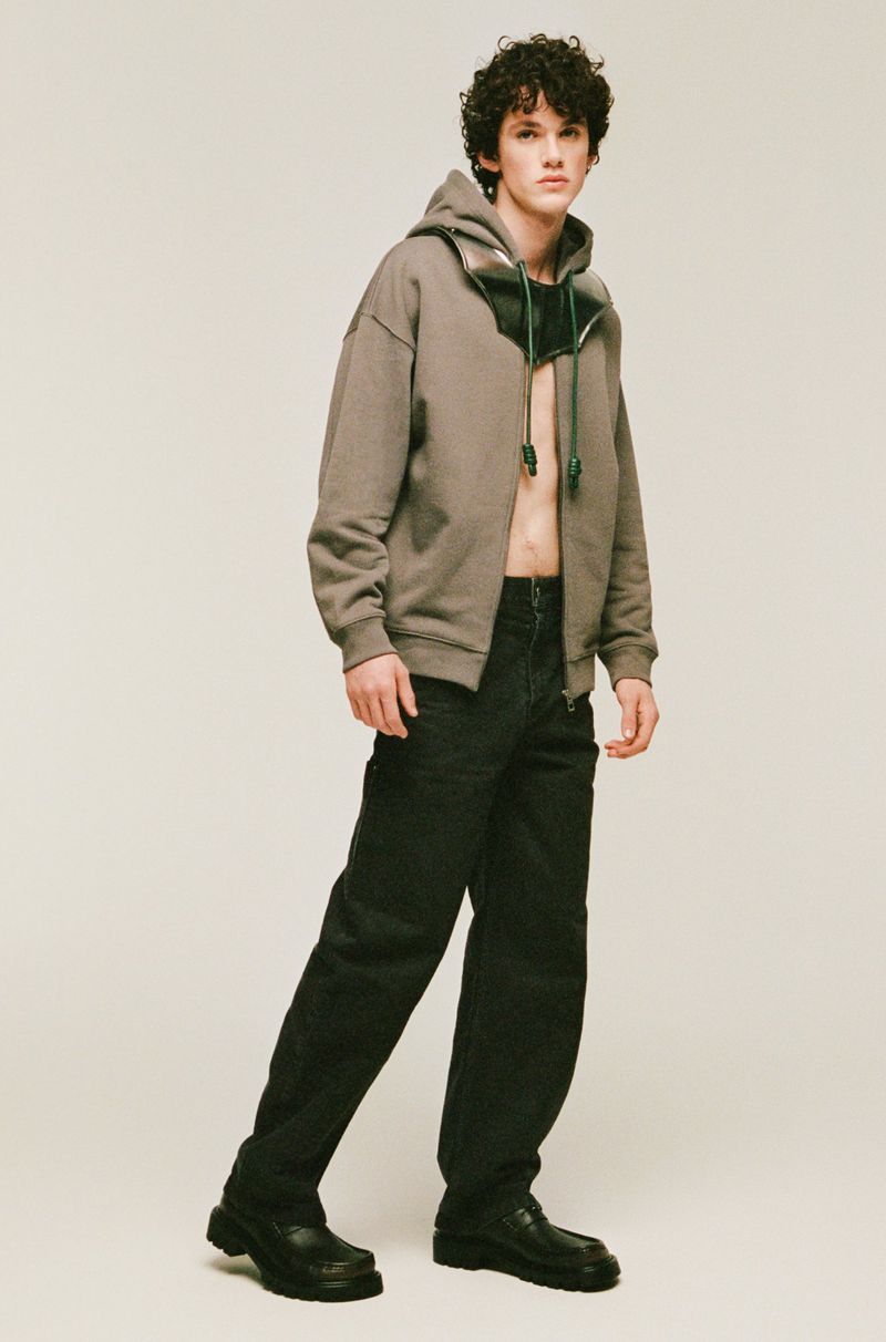 A young adult male with curly hair stands in a photo studio wearing a brown hoodie, black jeans, and black shoes in a portrait shot.