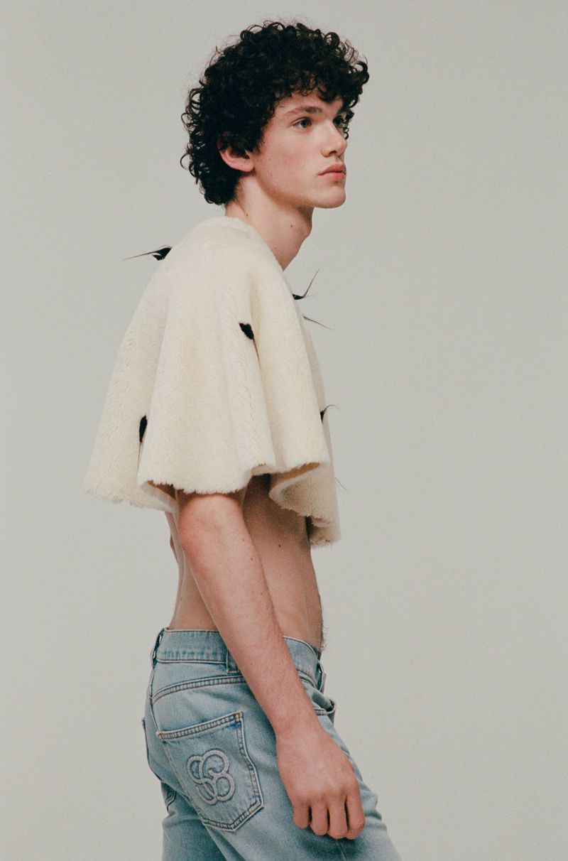 Young adult male poses in a studio wearing jeans and a cropped knit shirt. He has curly dark hair and pale skin. Studio lighting.