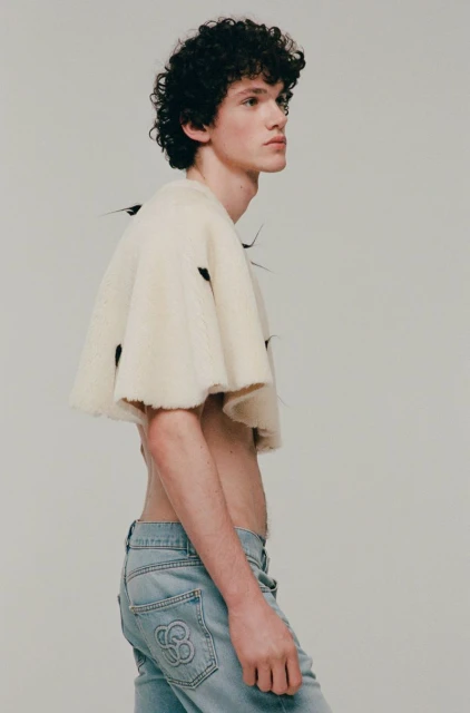 Young adult male poses in a studio wearing jeans and a cropped knit shirt. He has curly dark hair and pale skin. Studio lighting.