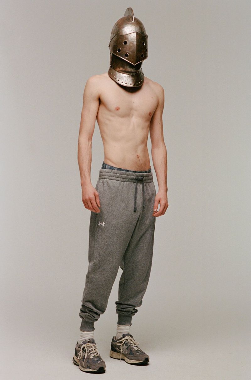 Man in sweatpants stands in a studio wearing a knight's helmet. He is shirtless and wearing grey New Balance sneakers. 