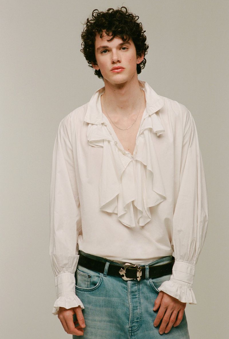 Studio shot of a young adult male model with curly hair wearing a white ruffled shirt, jeans, and a black belt posing in a photo studio.