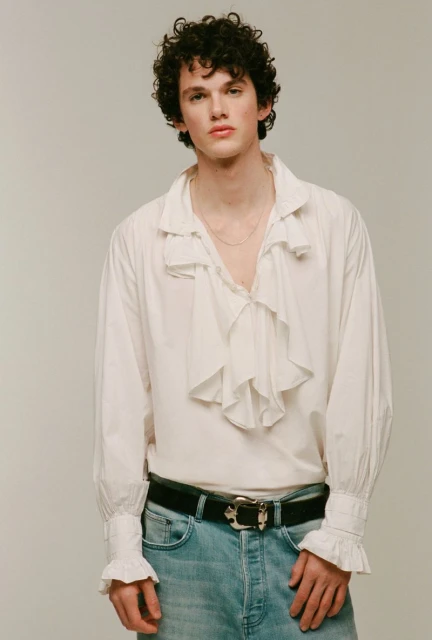 Studio shot of a young adult male model with curly hair wearing a white ruffled shirt, jeans, and a black belt posing in a photo studio.