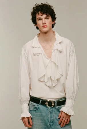 Studio shot of a young adult male model with curly hair wearing a white ruffled shirt, jeans, and a black belt posing in a photo studio.