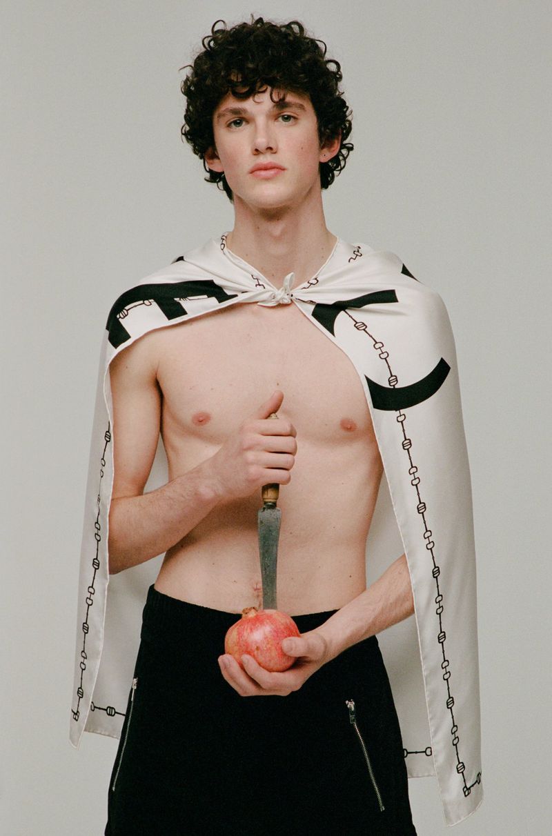 Young, shirtless man with cape poses indoors holding pomegranate and knife, set against a plain light background in a studio shot.