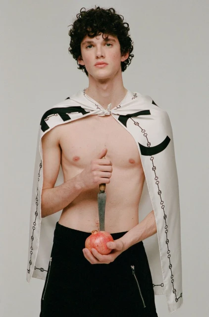 Young, shirtless man with cape poses indoors holding pomegranate and knife, set against a plain light background in a studio shot.