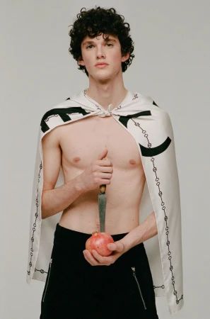 Young, shirtless man with cape poses indoors holding pomegranate and knife, set against a plain light background in a studio shot.
