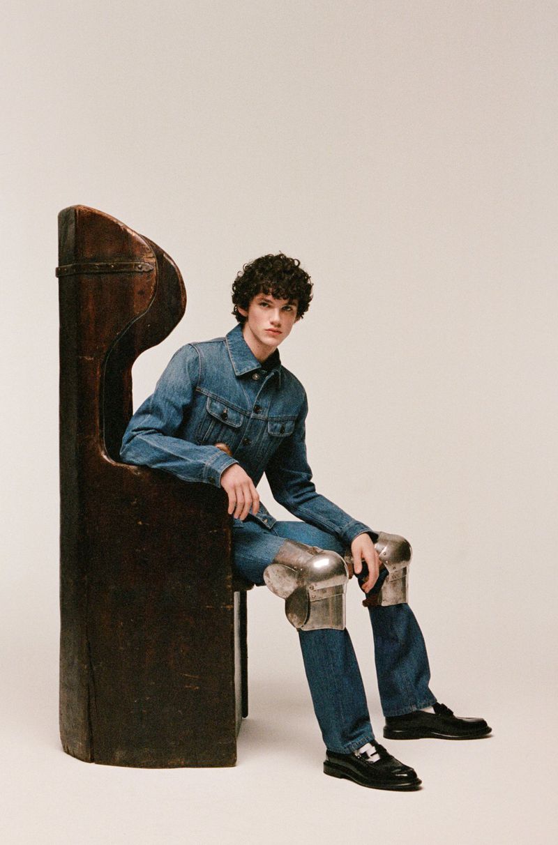 Adult male fashion model poses in a denim jacket and jeans, seated in a wooden chair with silver knee pads in a neutral indoor setting.
