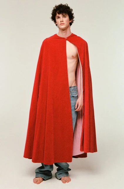 Male model with curly hair poses in studio wearing a red hooded cape and blue jeans. Barefoot, the model is centered on a white background.