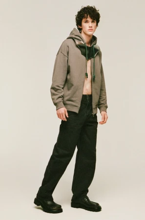 Full-length portrait of a male model wearing an olive green zip-up hoodie and dark trousers against a neutral studio backdrop.