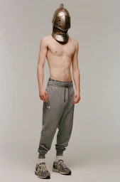 Shirtless male model wearing a metal knight helmet and grey sweatpants against a plain backdrop