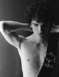 Portrait of a shirtless young man with curly hair and a decorative strap across his shoulder, shot in monochrome.