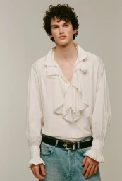 A young male model with dark curly hair wearing a white ruffled shirt and blue denim jeans against a solid neutral background.