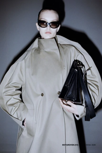 Fashion shot of a model wearing a tan trench coat and sunglasses, holding a black satchel handbag, against a grey background.