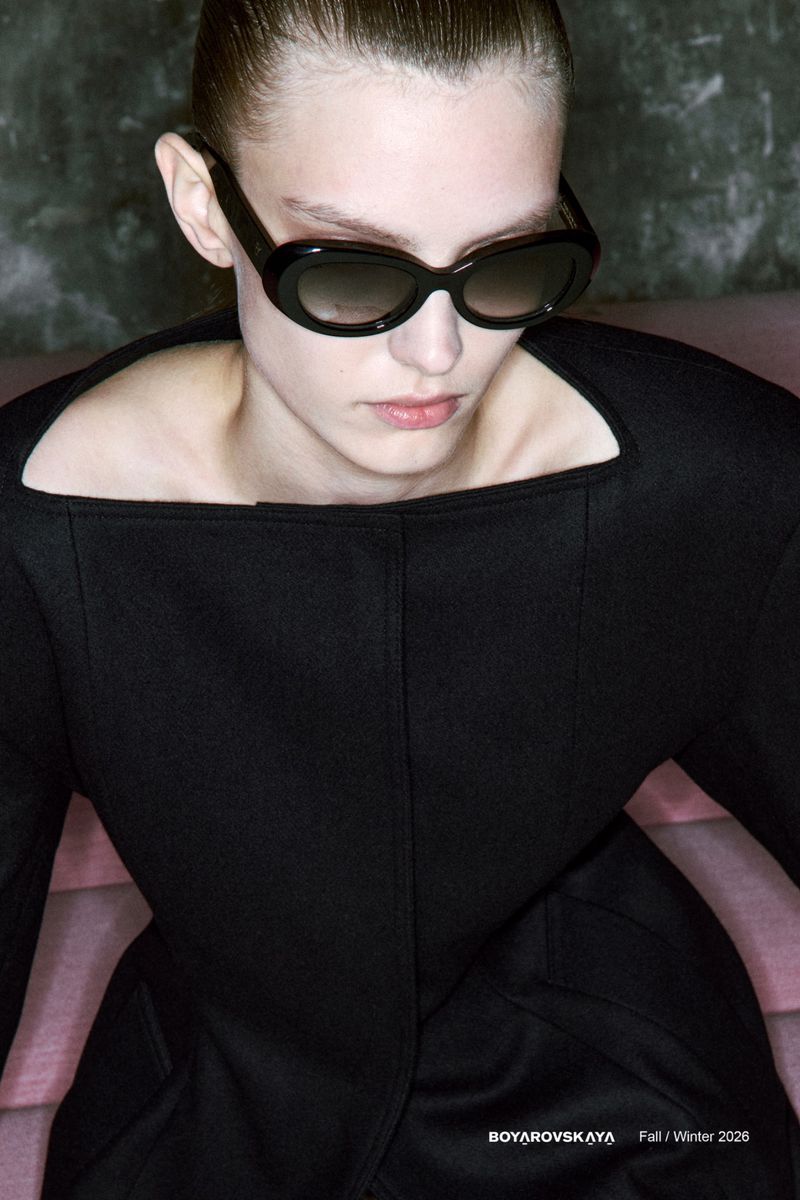 A fashionable woman with fair skin and dark sunglasses, wearing a black dress, captured in a studio with a muted pink and grey backdrop.