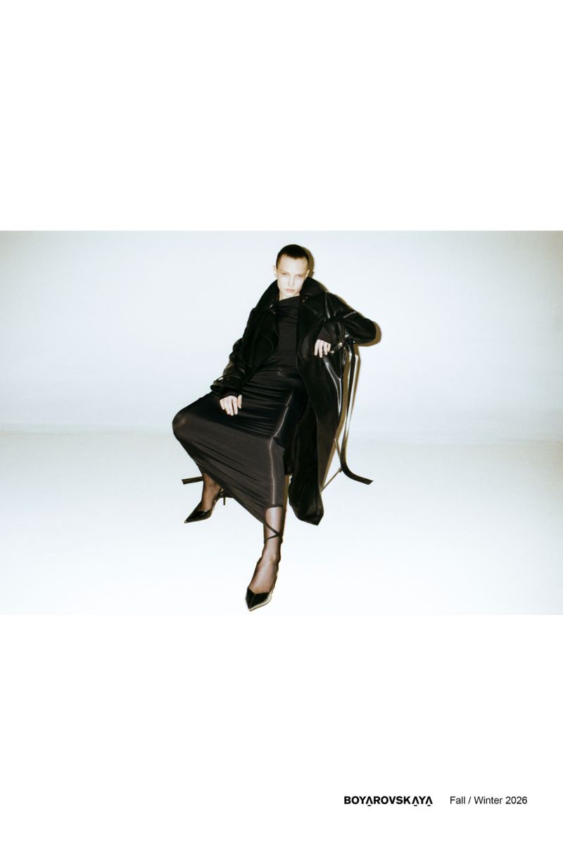 A slender adult woman is seated on a chair in a studio setting, wearing a black coat and skirt with sheer black tights and pointed heels.