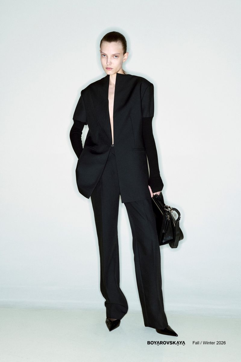 Fashion shot of a woman in a black jacket and pants, holding a purse, set against a white background in a studio environment.