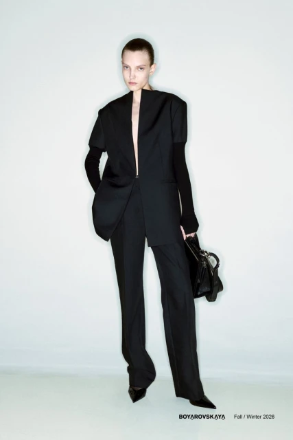 Fashion shot of a woman in a black jacket and pants, holding a purse, set against a white background in a studio environment.