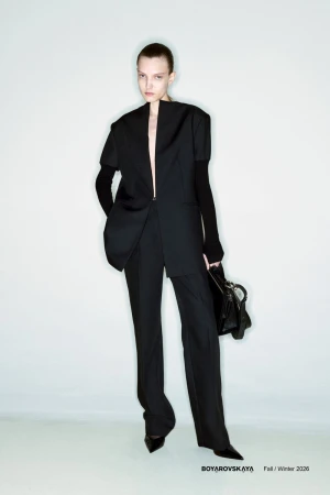 Fashion shot of a woman in a black jacket and pants, holding a purse, set against a white background in a studio environment.