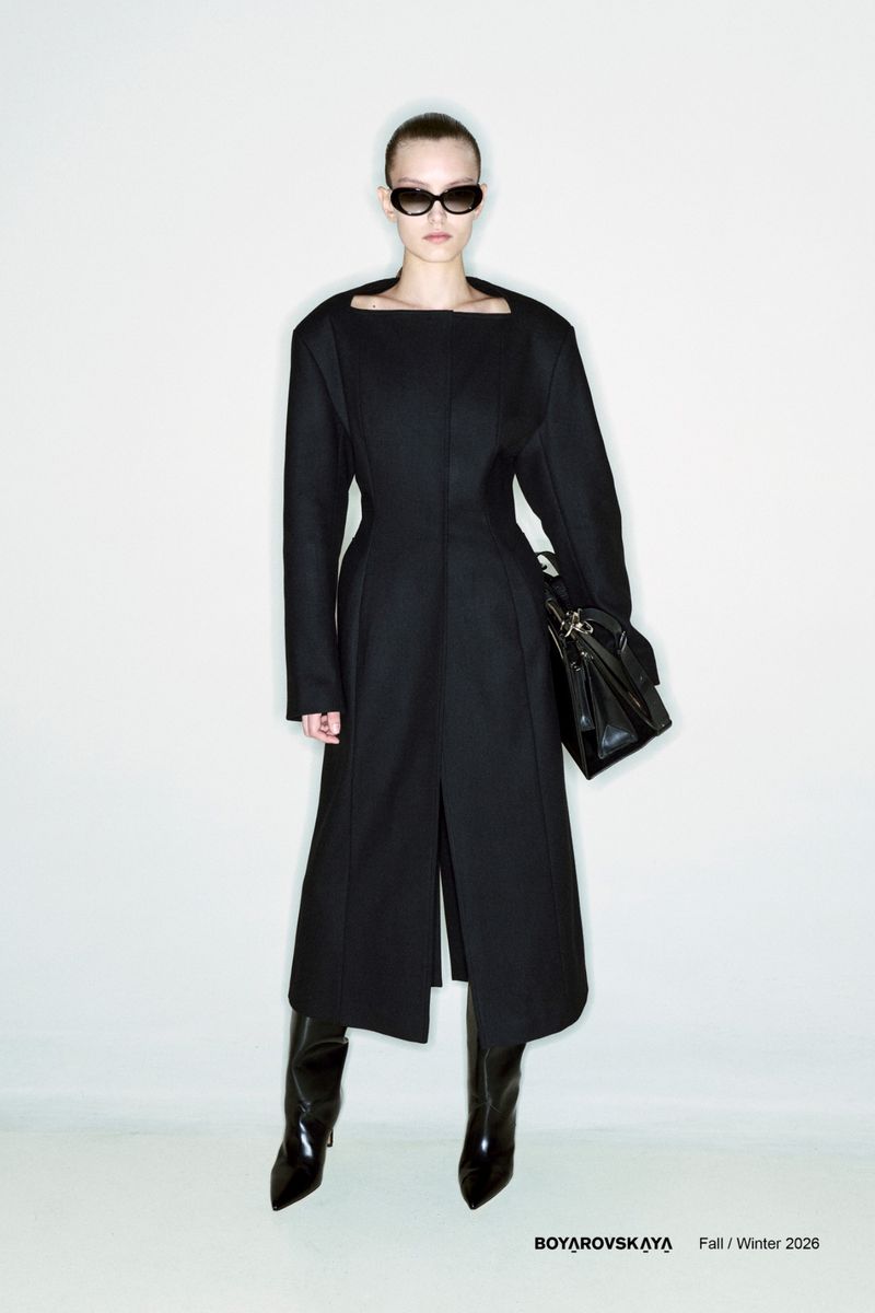 Model wearing a black structured coat and dark sunglasses holding a black handbag against a plain light grey studio background.