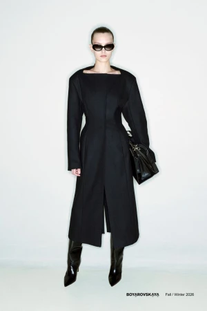 Model wearing a black structured coat and dark sunglasses holding a black handbag against a plain light grey studio background.