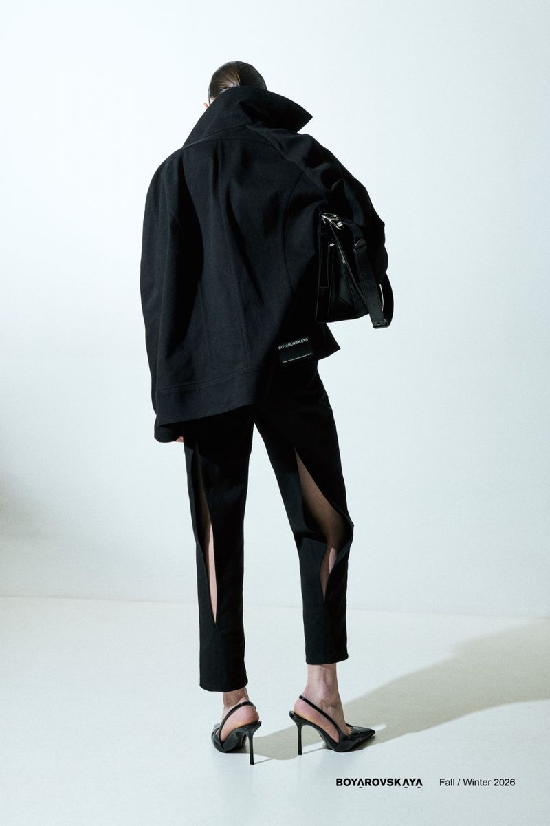 Fashion model poses in all-black designer ensemble. The back of the figure is to the camera in a minimalist studio environment.